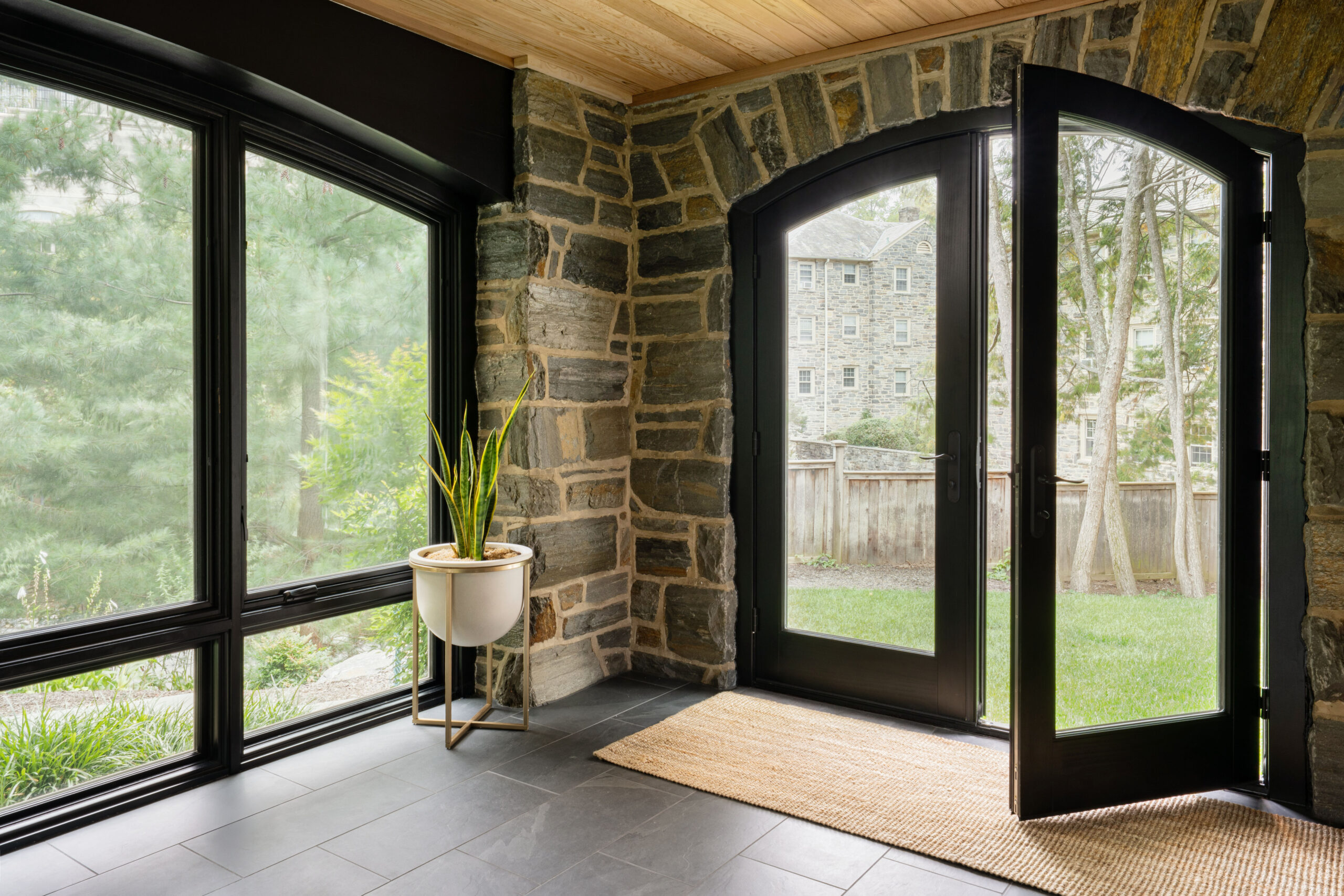All-season sunroom with stone walls, black-framed Pella Reserve Traditional windows, and garden views connecting indoor and outdoor living.