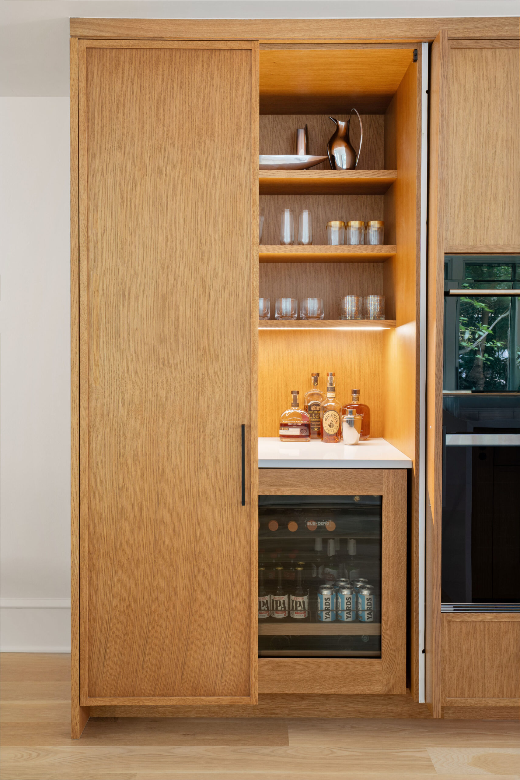 Custom white oak cabinetry with hidden bar featuring built-in shelving, glassware storage, and integrated beverage fridge.