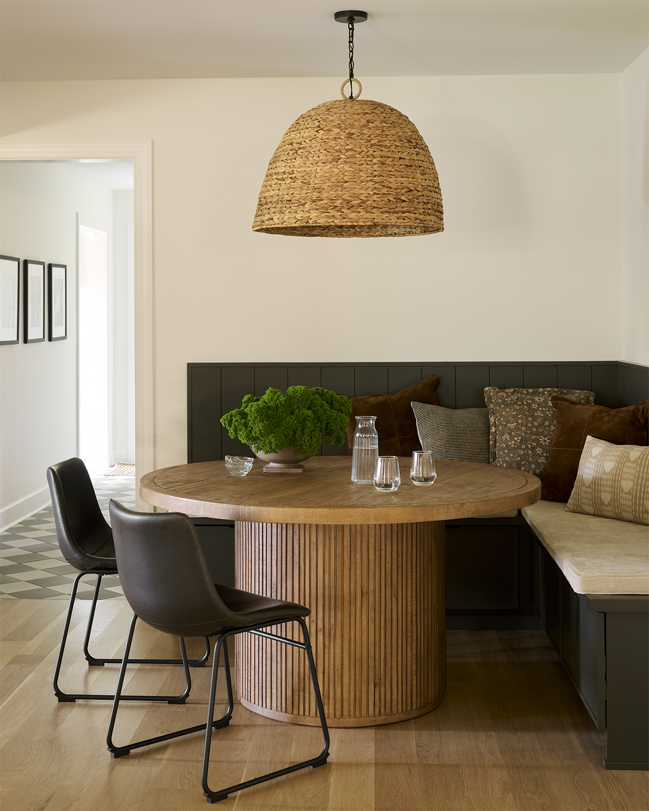 Breakfast nook with a round fluted wood pedestal table, dark paneled banquette seating, upholstered cushions, woven pendant light, and natural wood flooring.