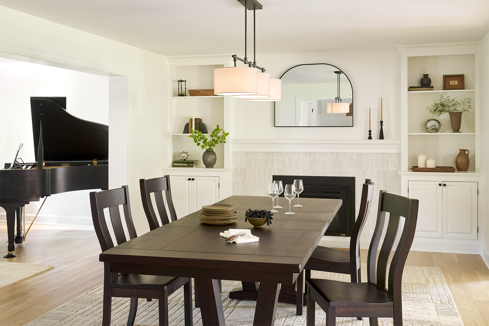 Reconfigured dining room with dark wood dining table, white built-in cabinetry, tiled fireplace, arched mirror, and adjoining piano room.