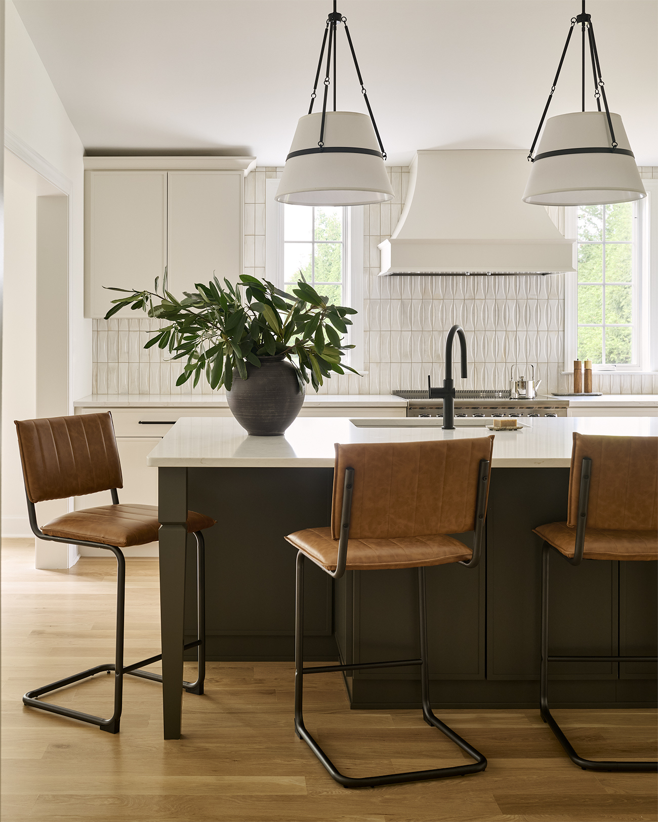 Chesterbrook kitchen renovation featuring a dark contrasting island, tan leather seating, oversized pendant lights, custom range hood, and textured white tile backsplash.