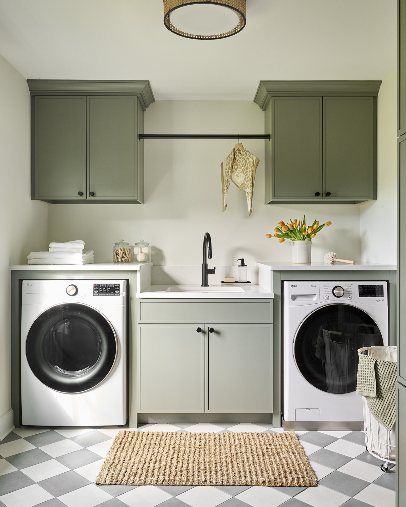 Chesterbrook laundry room featuring sage green cabinetry, side-by-side washer and dryer, central utility sink, matte black faucet, and gray-and-white checkerboard tile flooring.