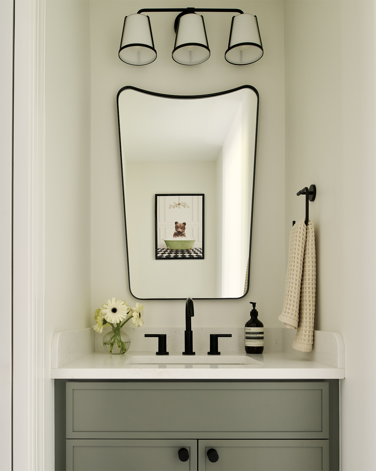 Powder room with sage green vanity, white countertop and integrated sink, matte black faucet, curved rectangular mirror, and three-light wall sconce.